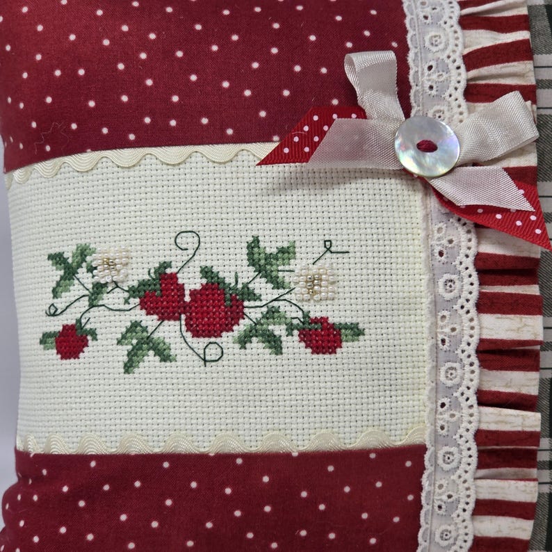 May include: A cushion with a red and white polka dot pattern and a cross-stitched strawberry design. The cushion features a white lace trim, a ribbon, and a button closure. The colour scheme is red, white, and green.