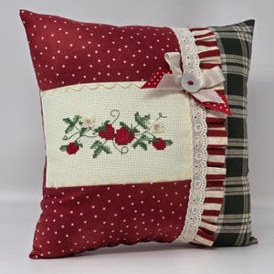 May include: A square cushion with a red polka dot pattern and a cross-stitched strawberry design. It has a white lace trim, a cream bow, and a plaid pattern on the back. The cushion is a decorative item for a home.