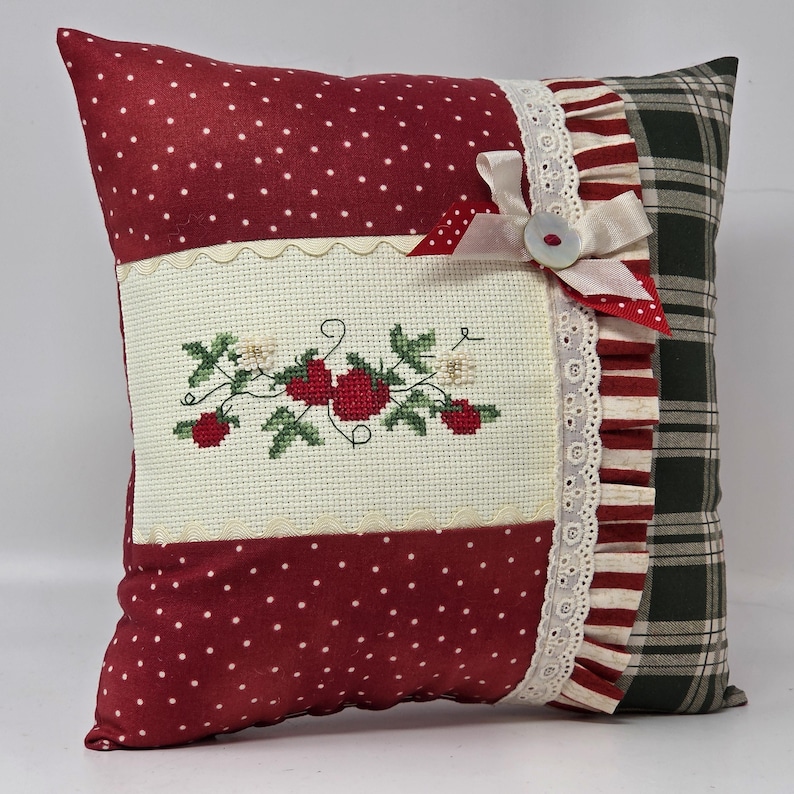 May include: A square cushion with a red polka dot pattern and a cross-stitched strawberry design. It has a white lace trim, a cream bow, and a plaid pattern on the back. The cushion is a decorative item for a home.