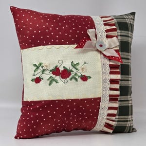 May include: A decorative cushion with a patchwork design. The front features red polka dot fabric, a cream embroidered panel with red strawberries, and a ruffled edge. The back is green plaid.