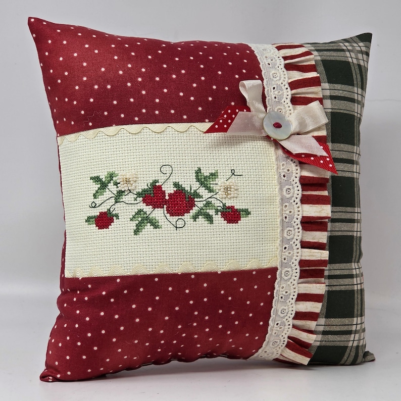 May include: A decorative cushion with a patchwork design. The front features red polka dot fabric, a cream embroidered panel with red strawberries, and a ruffled edge. The back is green plaid.