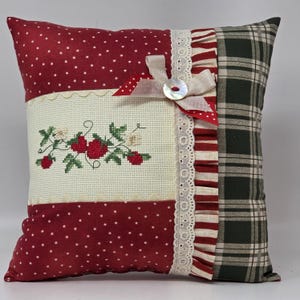 May include: A square cushion with a patchwork design. It features red polka dot fabric, a cross-stitched floral design, and a green plaid pattern. The cushion is embellished with lace, a ribbon, and a button.