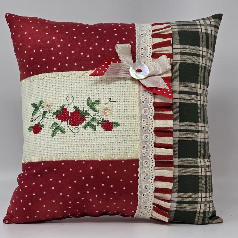 May include: A square cushion with a patchwork design. It features red polka dot fabric, a cross-stitched floral design, and a green plaid pattern. The cushion is embellished with lace, a ribbon, and a button.