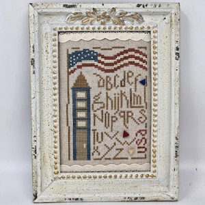 May include: A framed cross-stitch sampler on beige fabric featuring a waving American flag, a tall blue and brown tower, and the alphabet in brown thread. The text USA is stitched in red, and the white wooden frame has a distressed finish.