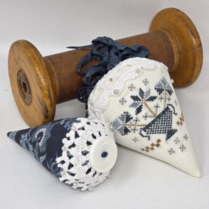May include: Two cone-shaped fabric ornaments with white lace trim and pearl accents. One is navy with a floral pattern, the other is off-white with a cross-stitch design. A wooden spool is in the background.