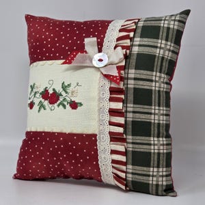 May include: Square cushion with a patchwork design. It features a red polka dot pattern, a cross-stitched strawberry design, and a green and white plaid pattern. A white ribbon and button add a decorative touch.