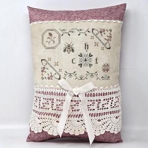May include: A decorative pillow with a floral and bee cross-stitch design. The pillow features a cream-colored center with embroidered details, a lace trim, and a mauve fabric border. A white ribbon with a heart-shaped button adds a delicate touch.