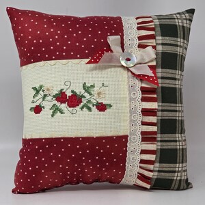 May include: Square cushion with a patchwork design. It features red fabric with white polka dots, a cross-stitched floral design, and a green plaid pattern. A white ribbon and button accent the cushion.