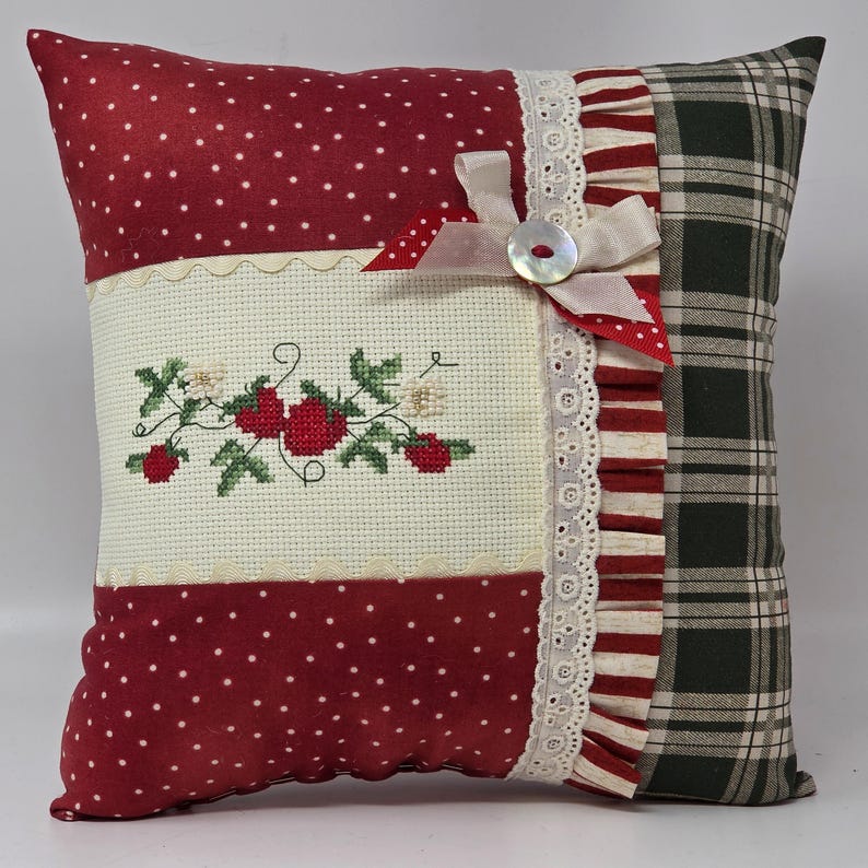 May include: Square cushion with a patchwork design. It features red fabric with white polka dots, a cross-stitched floral design, and a green plaid pattern. A white ribbon and button accent the cushion.