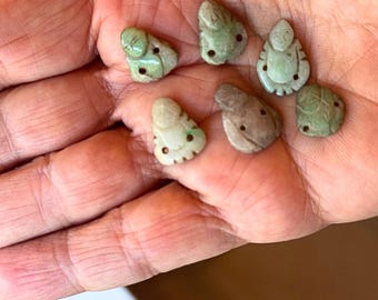 May include: Six small jade green and brown carved pendants in the shape of a person. The pendants have two holes at the top for stringing.