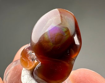Puede incluir: Una piedra de ágata pulida e iridiscente con un color rojo intenso, marrón y morado. La piedra tiene una forma única y orgánica y se muestra sobre una base blanca.