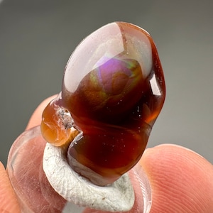 May include: A polished, iridescent agate stone with a deep red, brown, and purple color. The stone has a unique, organic shape and is displayed on a white base.