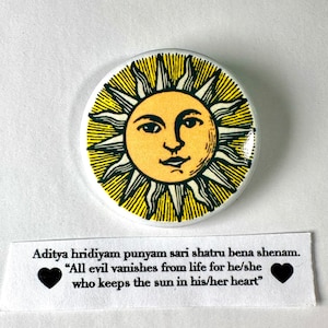 May include: A yellow and black sun with a face on it. The sun is on a white button with a black border. The text on the button reads "Aditya hridiyam punyam sari shatru bena shenam. "All evil vanishes from life for he/she who keeps the sun in his/her heart""