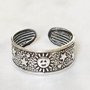 May include: An open-band silver ring featuring a textured background with embossed sun and star motifs. The metal has an oxidized finish in the recessed areas to highlight the celestial design, and the interior band shows horizontal line detailing.