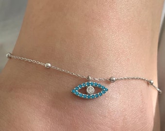 Könnte beinhalten: Ein silbernes Fußkettchen mit einem blauen bösen Blick-Anhänger. Der Anhänger hat einen kleinen, klaren Stein in der Mitte.