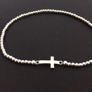 May include: A silver beaded bracelet with a small cross pendant. The bracelet is made of small, round beads strung together, with a simple cross charm centered on the band. The jewelry is displayed against a dark background.
