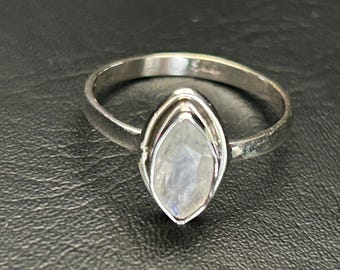 May include: A silver ring featuring a marquise-cut moonstone with an iridescent sheen. The gemstone is set in a double bezel. The ring has a simple, rounded band. The ring is shown on a dark, textured surface.