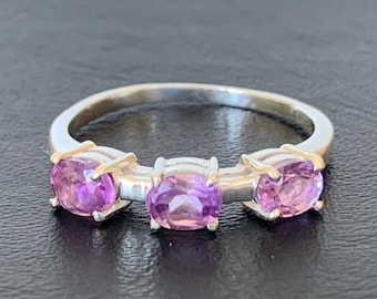May include: A silver ring featuring three oval-cut, vibrant purple gemstones. The gemstones are set in a row, creating a classic and elegant design. The ring is displayed on a dark surface, highlighting the sparkle of the stones.