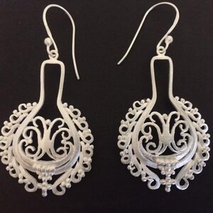 Può includere: Un paio di orecchini in filigrana d'argento con un design delicato e intricato. Gli orecchini hanno la forma di una lacrima con un motivo circolare traforato.