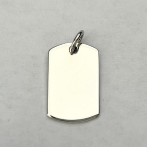 Pode incluir: Um pingente de placa de identificação para cães, de cor prateada polida, com uma base arredondada e uma pequena argola na parte superior para uma corrente. A placa está em branco, oferecendo espaço para gravação ou personalização.