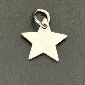 May include: A silver star charm with a loop for attaching to a necklace or bracelet. The charm is a five-pointed star with a polished finish. The background is a dark grey surface.