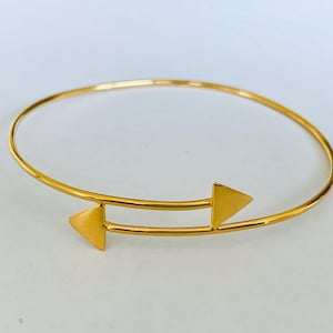 May include: A gold-coloured bangle bracelet with an arrow design. The bracelet has a circular band with two arrow-shaped accents, each with a triangular head. The jewellery is displayed on a white surface.