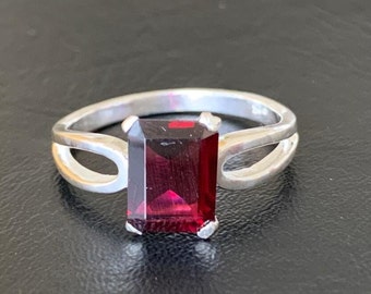 Könnte beinhalten: Ein silberner Ring mit einem dunkelroten, smaragdgeschliffenen Edelstein.