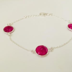 Op de afbeelding: Een zilveren armband met vier grote, ronde, fuchsia-kleurige edelstenen. De edelstenen zijn gezet in zilveren bezels en verbonden door een fijne zilveren ketting. De armband heeft een karabijnsluiting.