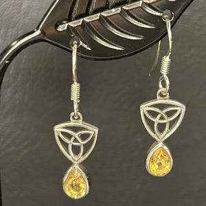 May include: Silver-toned dangle earrings featuring a Celtic knot design within a triangular frame, with a teardrop-shaped, yellow gemstone at the bottom. The earrings are displayed on a black stand.