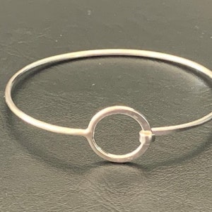 Puede incluir: Un brazalete de plata con un dije circular. El dije circular está abierto y tiene una pequeña abertura en la parte superior.