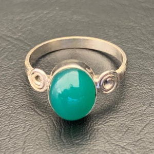 Puede incluir: Un anillo de plata con una piedra grande, ovalada y verde brillante. El anillo tiene una banda simple y detalles decorativos en espiral a cada lado de la piedra. El anillo está sobre un fondo oscuro.
