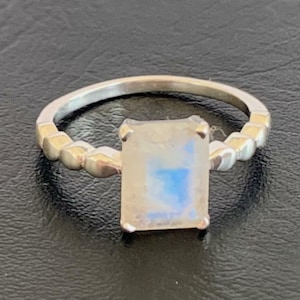 Puede incluir: Un anillo de plata con una piedra de luna rectangular engastada en un engaste de bisel. La banda está decorada con una fila de pequeñas cuentas.