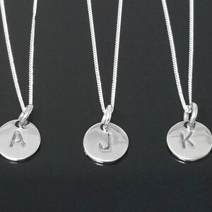 Op de afbeelding: Drie zilveren kettingen met ronde hangers met de letters A, J en K.