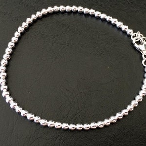 May include: A silver beaded bracelet with a lobster clasp and a small silver charm. The bracelet is made of small, round beads.