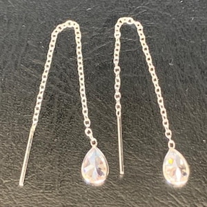 Puede incluir: Un par de pendientes de hilo plateados. Cada pendiente presenta una delicada cadena que conduce a un cristal transparente en forma de lágrima. Los pendientes tienen un diseño simple y elegante.