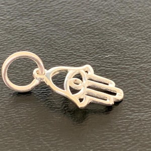 May include: A silver charm in the shape of a Hamsa hand with an eye in the center. The charm has a loop at the top for attaching to a bracelet or necklace.