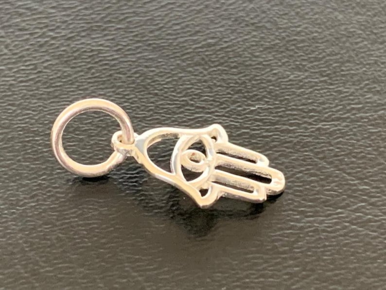 May include: A silver charm in the shape of a Hamsa hand with an eye in the center. The charm has a loop at the top for attaching to a bracelet or necklace.