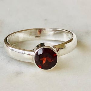 May include: A silver ring featuring a round, deep red gemstone. The ring has a polished band and a bezel setting. The gemstone is the focal point. The ring is a classic design, suitable for everyday wear.