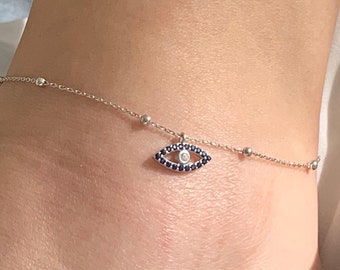 Könnte beinhalten: Ein silbernes Fußkettchen mit einem blauen bösen Blick-Anhänger. Der Anhänger hat einen weißen Stein in der Mitte.
