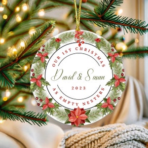 May include: A round Christmas ornament with a gold string for hanging. The ornament features a wreath of green pine needles, red berries, and poinsettias. Text reads "Our 1st Christmas" and "David & Susan 2023 As Empty Nesters."