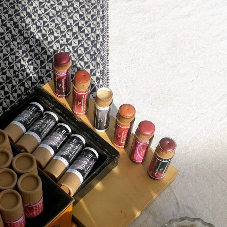 Puede incluir: Una colecci&oacute;n de tubos de b&aacute;lsamo labial en varios colores, incluyendo rojo, rosa y naranja. Los tubos est&aacute;n hechos de cart&oacute;n y tienen una etiqueta negra con el texto "Lipbalm All Australian" impreso en ellos.