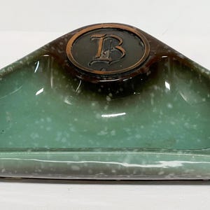 May include: Triangular ashtray with a speckled turquoise and brown glaze. A bronze-colored medallion with the letter "B" is centered on the ashtray. The ashtray has a shallow, curved design with a built-in rest for cigarettes.