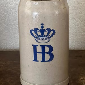 Könnte beinhalten: Ein zylindrischer, cremefarbener Bierkrug aus Keramik mit einem blauen Kronen-Emblem und den Buchstaben "HB". Der Krug hat eine leicht strukturierte Oberfläche und einen abgerundeten Boden. Das Design ist schlicht und klassisch.