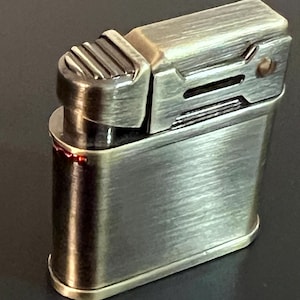 May include: A vintage-style metal lighter with a brushed finish. The lighter has a rectangular body with rounded edges and a textured top for ignition. The color is a mix of bronze and silver, giving it a classic, antique appearance.