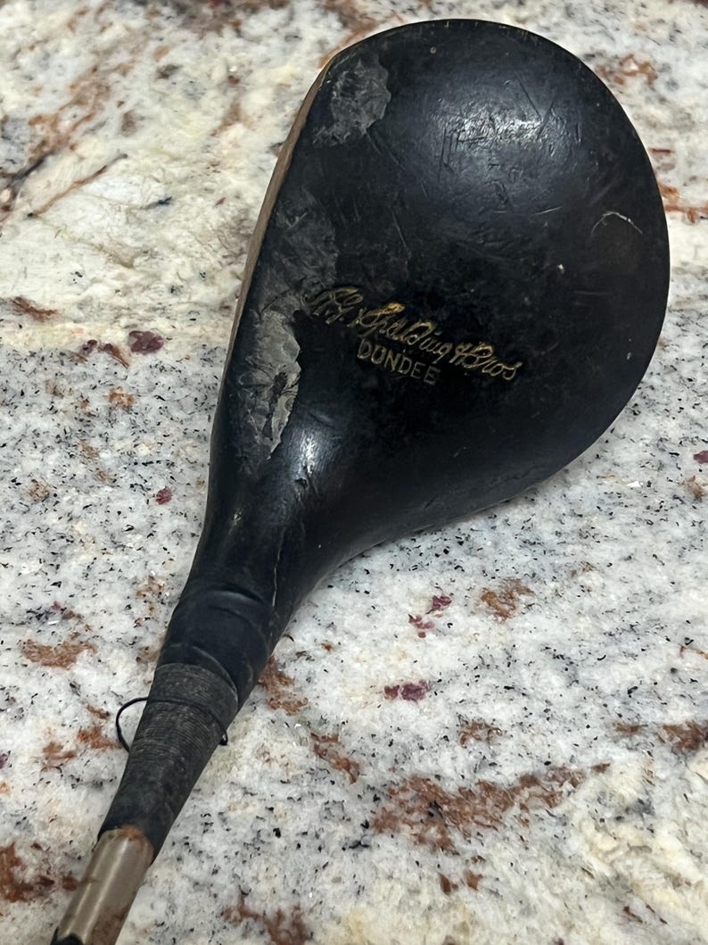Pu&ograve; includere: Un'antica mazza da golf con una testa in legno scuro e usurato e un lungo manico sottile. La testa della mazza presenta segni di usura visibili, con il testo "A.G. Spalding Bros. DUNDEE" inciso sulla superficie. L'impugnatura &egrave; avvolta con un'impugnatura scura.
