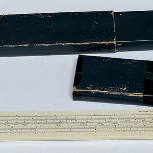 May include: A vintage slide rule with a cream-colored body and black markings, accompanied by two dark green cases. The slide rule features a metal frame and appears to be well-preserved, showcasing its age and use. The cases are rectangular and show signs of wear.