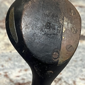 Pu&ograve; includere: Primo piano di una testa di mazza da golf in legno vintage. La testa della mazza &egrave; marrone scuro con usura visibile, con accenti metallici e viti. Il numero "2" &egrave; stampato sulla faccia della mazza. Un classico oggetto da collezione sportivo.