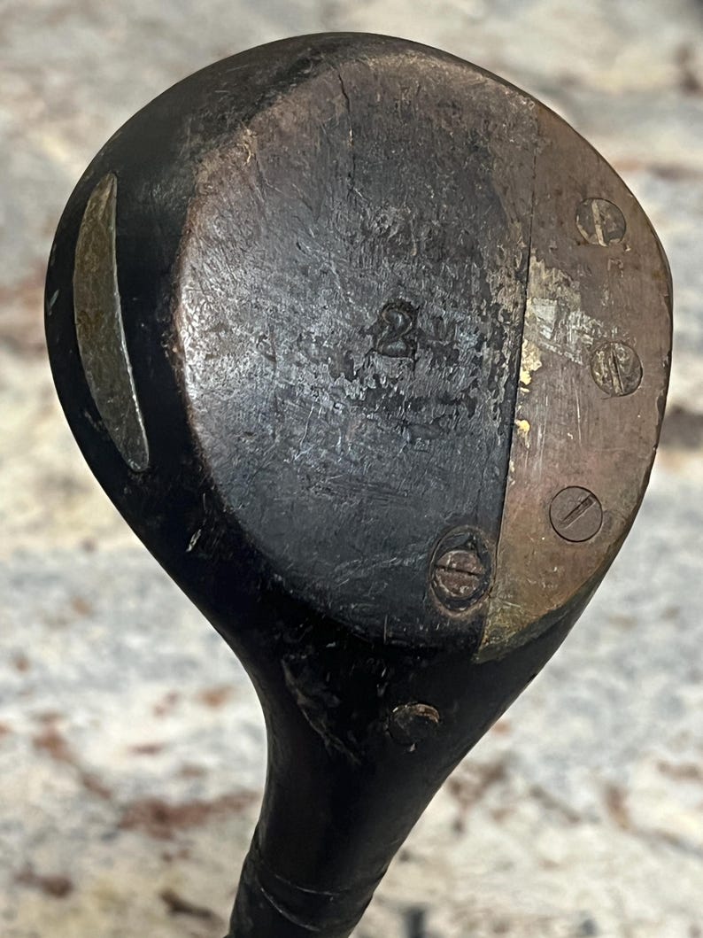 Pu&ograve; includere: Primo piano di una testa di mazza da golf in legno vintage. La testa della mazza &egrave; marrone scuro con usura visibile, con accenti metallici e viti. Il numero "2" &egrave; stampato sulla faccia della mazza. Un classico oggetto da collezione sportivo.