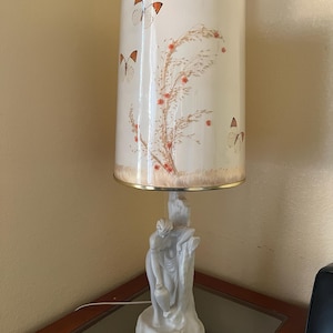 May include: A vintage table lamp with a white lampshade featuring butterflies and floral designs. The base is a white sculpture of two figures. The lampshade has a gold trim.