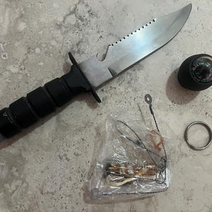 May include: A survival knife with a serrated blade, black handle, and a silver-colored blade. Includes a compass, a small metal ring, and a small survival kit in a clear plastic bag.